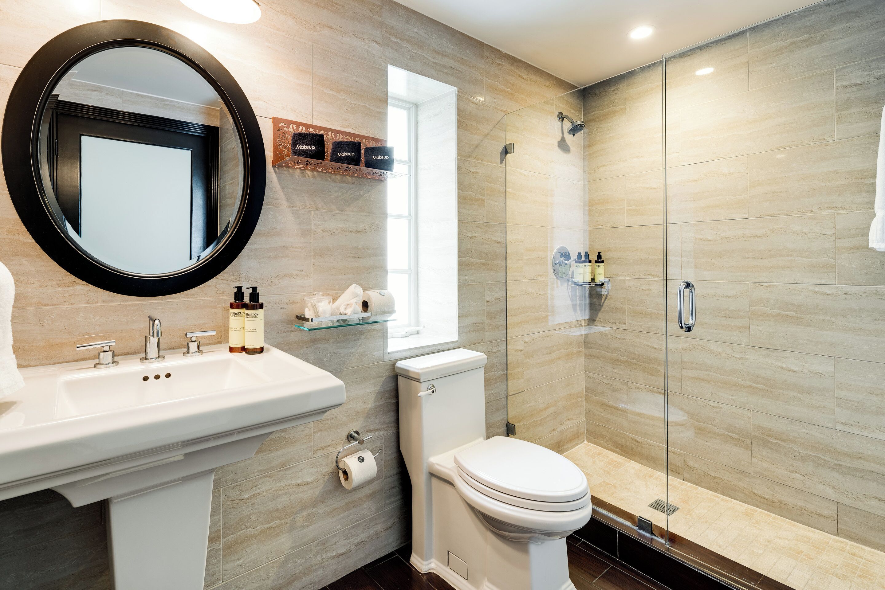 A modern bathroom with a round mirror, a sink with two faucets, a toilet, and a glass-enclosed shower area.
