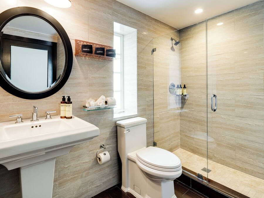 A modern bathroom with a round mirror, a sink with two faucets, a toilet, and a glass-enclosed shower area.