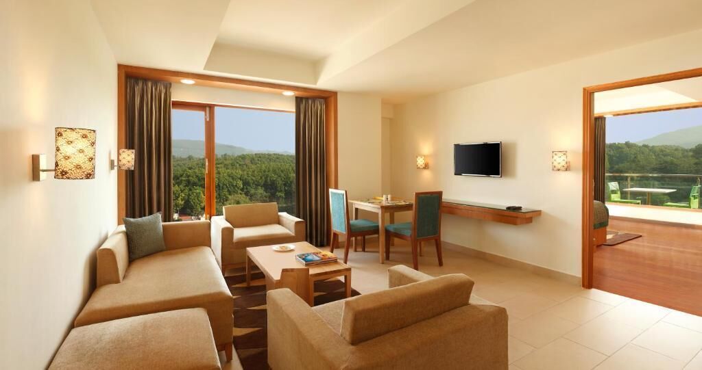 A spacious living room with a sofa set, a dining table, and chairs. There is a flat-screen TV mounted on the wall and a balcony with a view of the outdoors.