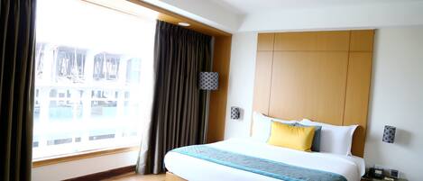 A hotel room with a large bed, a nightstand, a lamp, and a view of the city through the window.