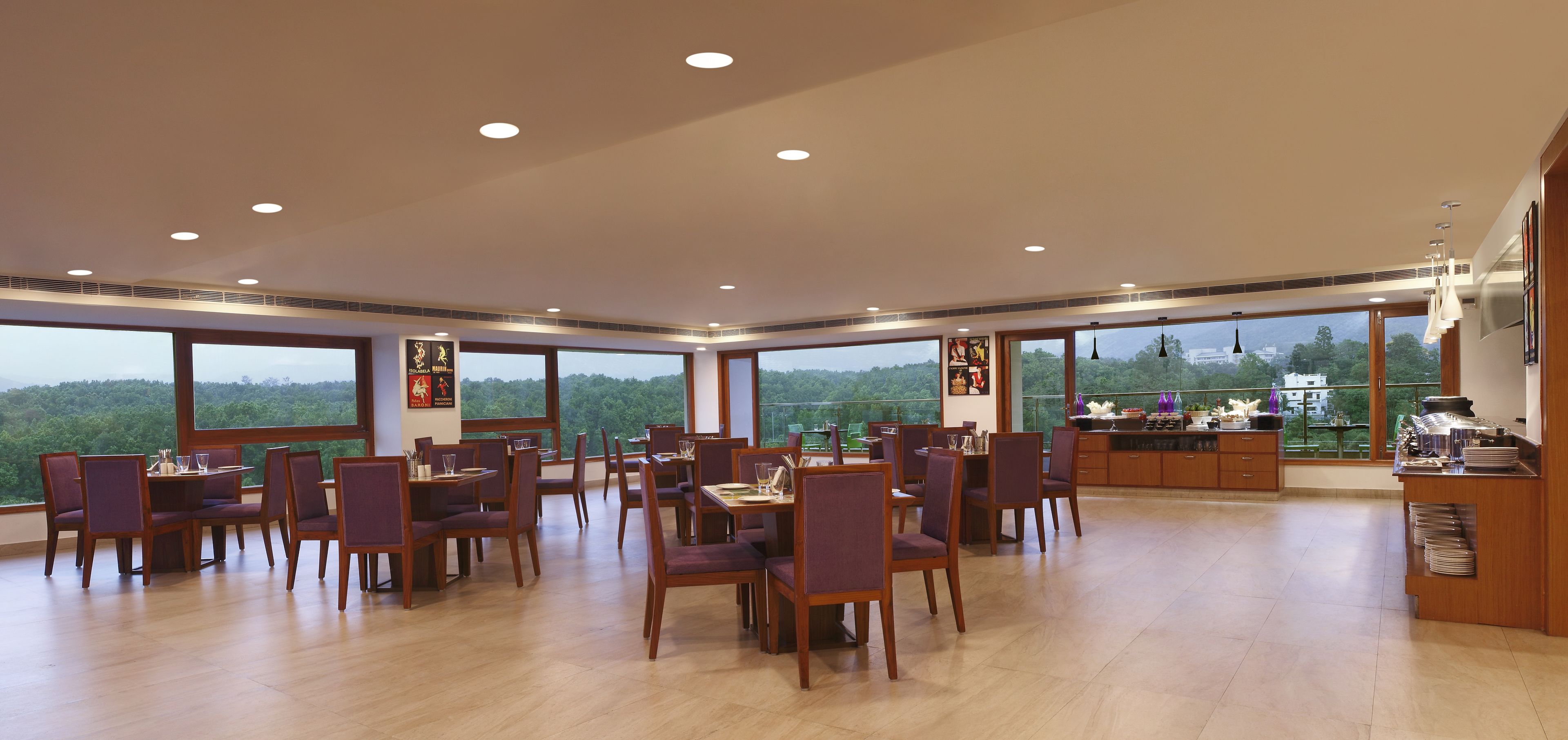 A spacious dining area with multiple tables and chairs, large windows offering a view of the greenery outside, and a kitchen area with modern appliances and utensils visible.