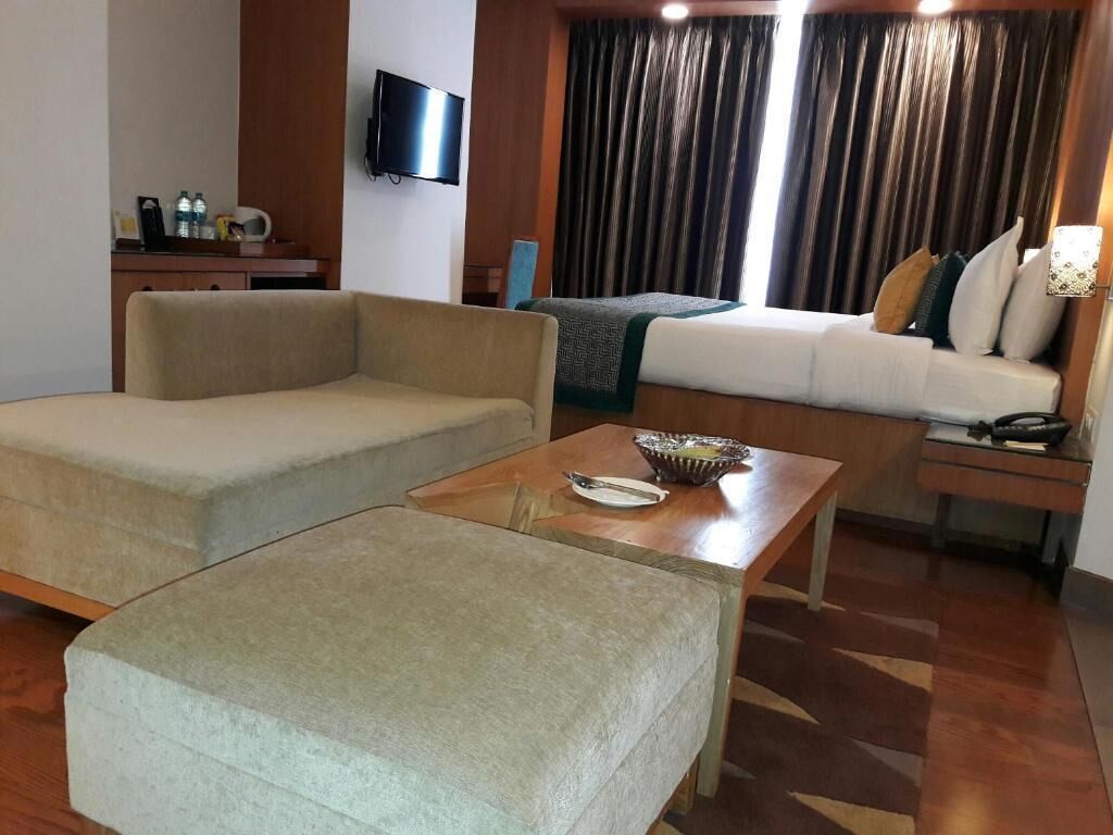 A hotel room with a sofa, a small table, a TV mounted on the wall, and a window with curtains.