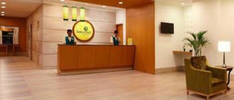 Zona de recepción con un mostrador de madera, dos empleados, un logotipo circular y un sillón verde.
