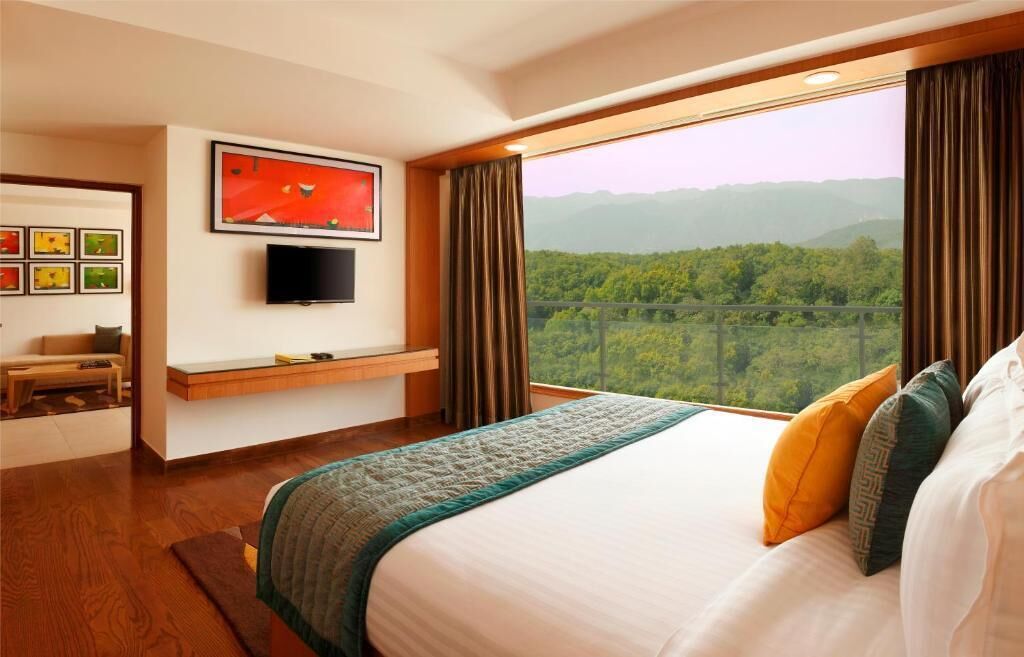 A hotel room with a large bed, a flat-screen TV mounted on the wall, a desk with a chair, and a window offering a view of a forested landscape.