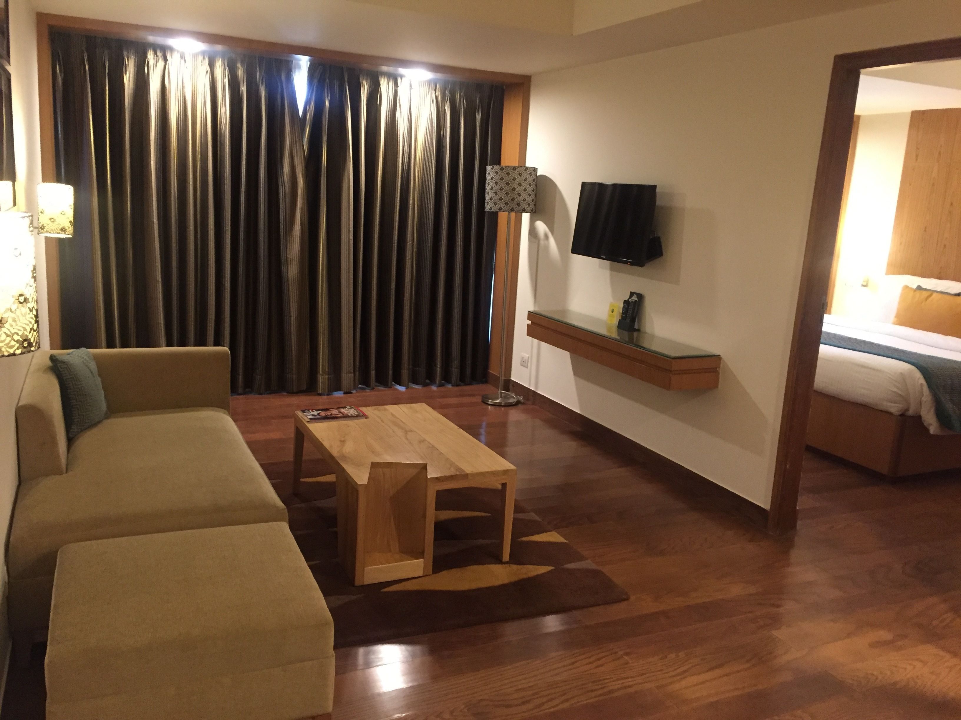 A living room with a sofa, a coffee table, a TV mounted on the wall, and a bed in the background.