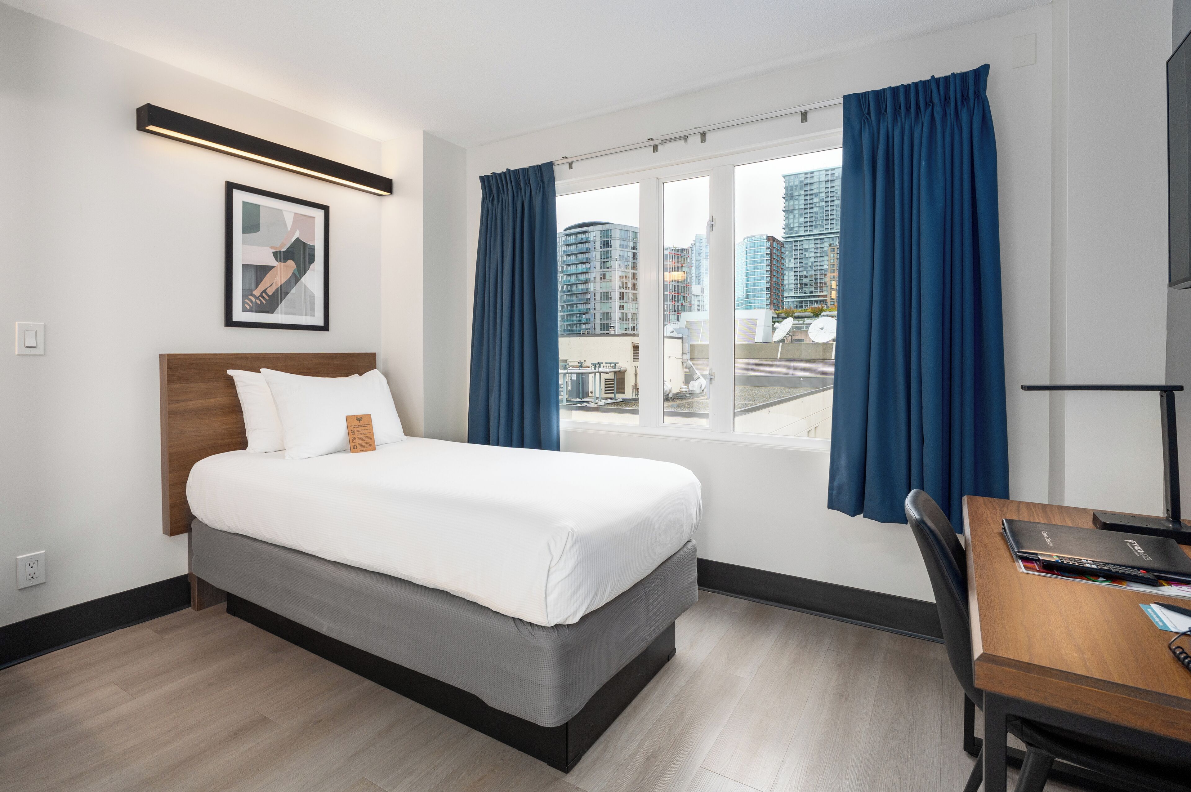 Quarto de hotel com cama, escrivaninha, cadeira, janela com cortinas, quadro na parede e vista para a cidade.