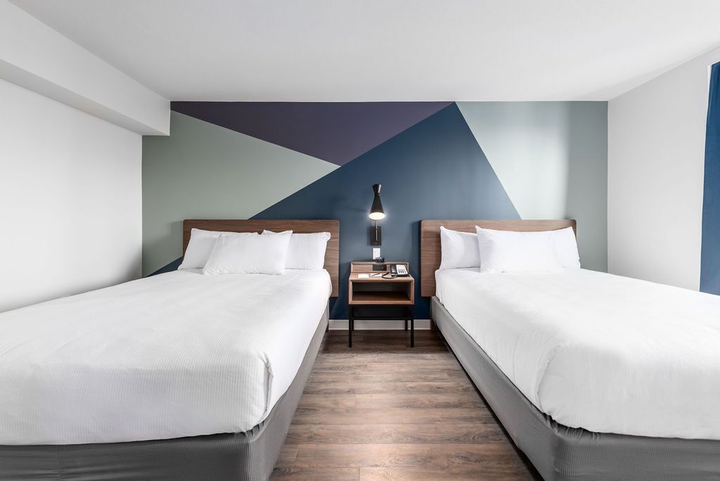 A hotel room with two beds, a nightstand with a lamp, and a geometric wall design.