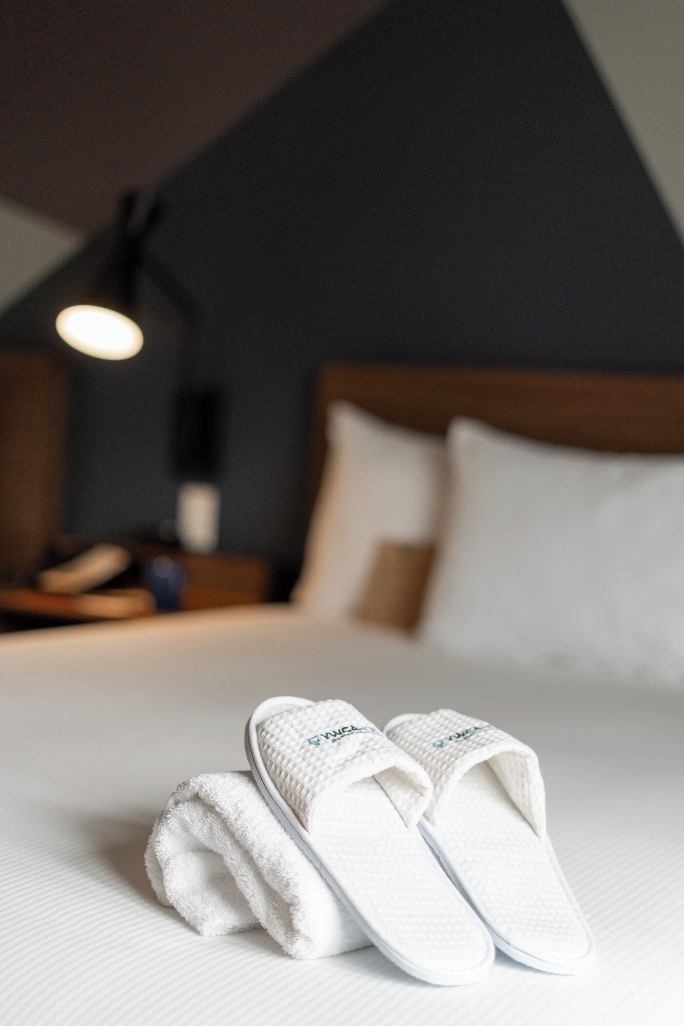 A neatly made bed with white linens and a pair of white slippers placed on it.