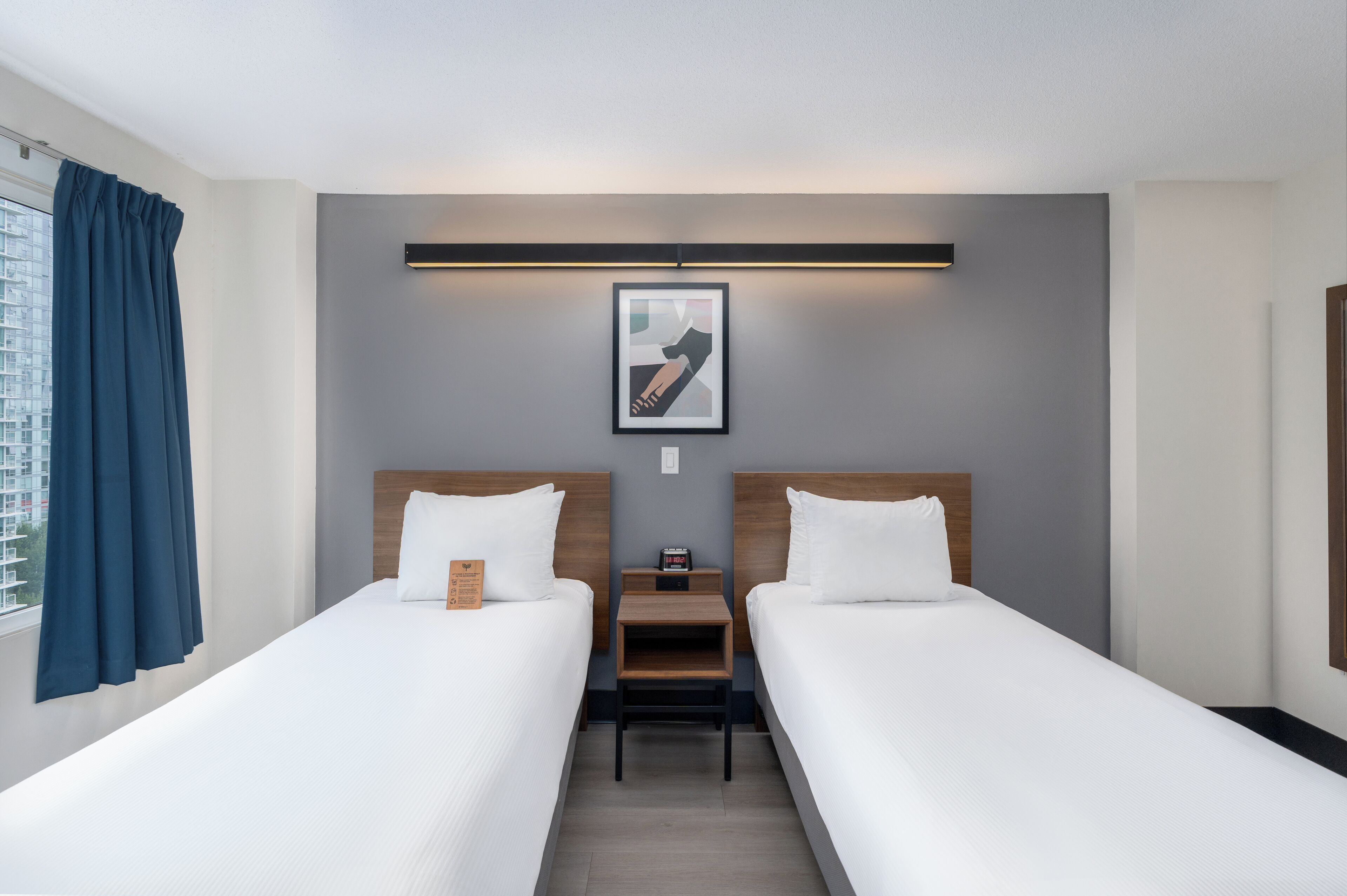 Une chambre d’hôtel avec deux lits simples, une table de chevet, un tableau encadré au mur et une fenêtre avec des rideaux.