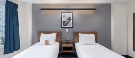 Une chambre d’hôtel avec deux lits simples, une table de chevet, un tableau encadré au mur et une fenêtre avec des rideaux.