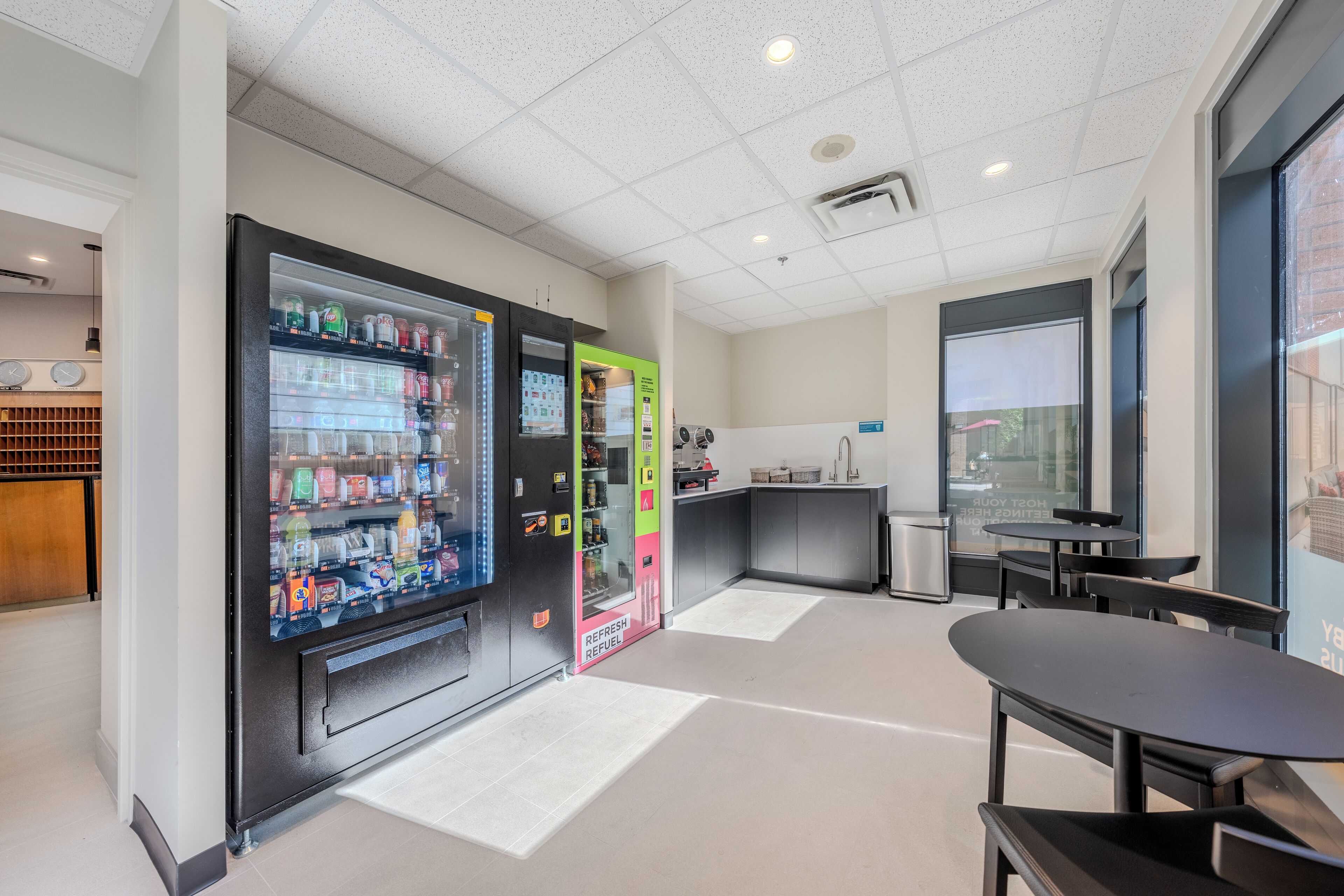 A modern hotel lobby with a vending machine, a refreshment station, and a seating area with tables and chairs.