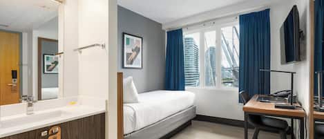 Une chambre d’hôtel moderne équipée d’un lit, d’un bureau, d’un ordinateur, d’un miroir, d’une fenêtre avec des rideaux et offrant une vue sur les bâtiments.