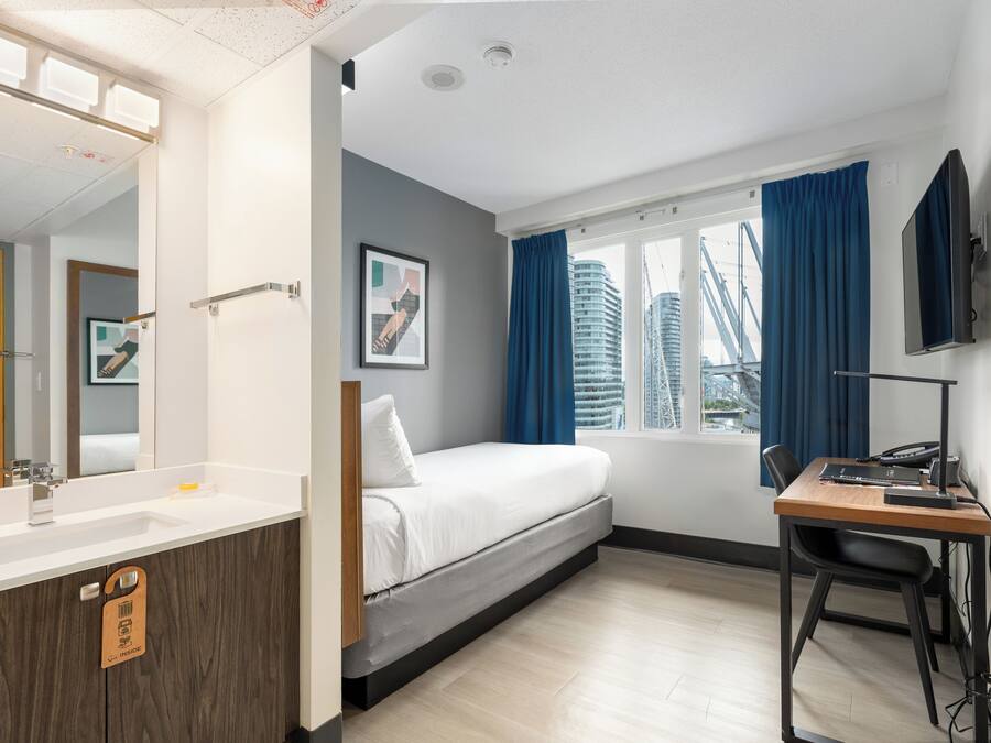 A modern hotel room with a bed, a desk, a computer, a mirror, a window with curtains, and a view of buildings.
