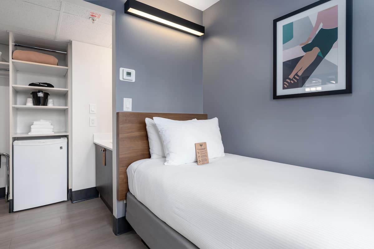 A modern hotel room with a bed, a mini-fridge, a built-in wardrobe, and a wall-mounted artwork.