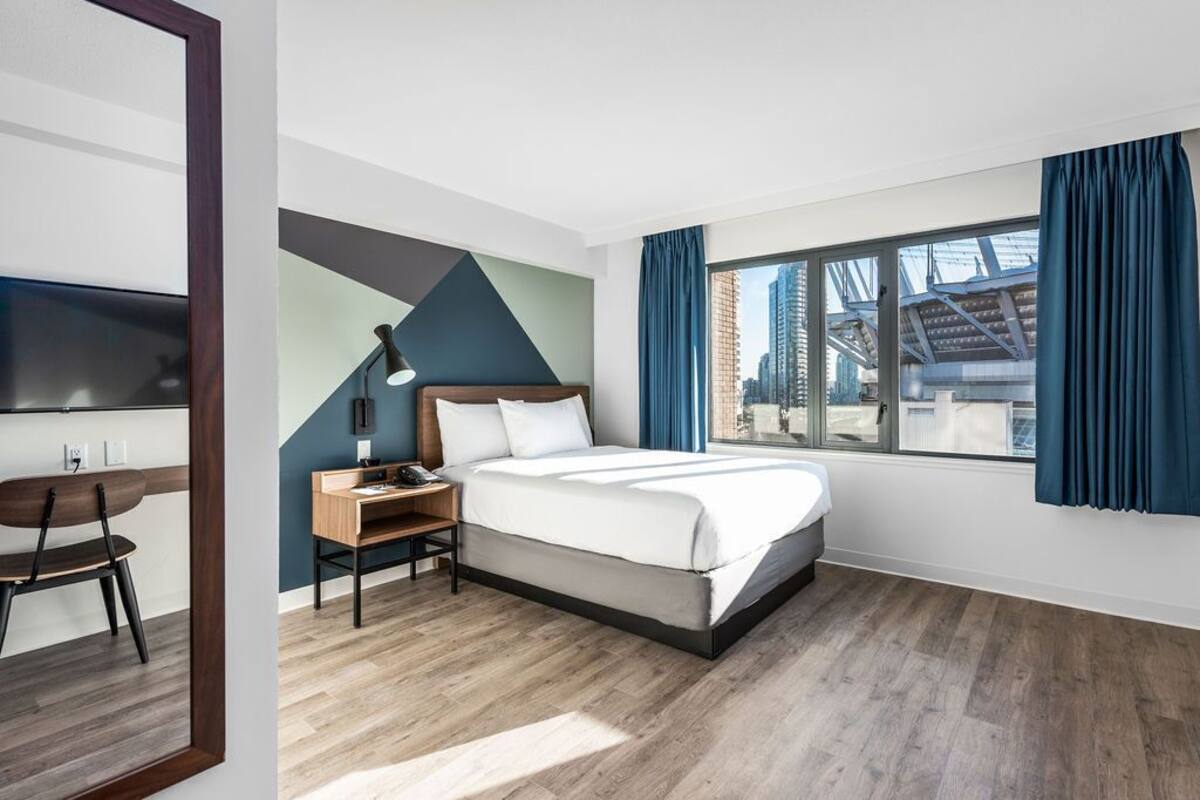 Une chambre d’hôtel moderne équipée d’un lit, d’un bureau, d’une chaise et de grandes fenêtres donnant sur la ville.