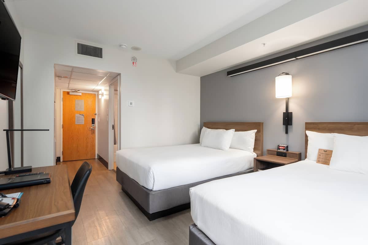 A hotel room with two beds, a desk, and a nightstand with a lamp.
