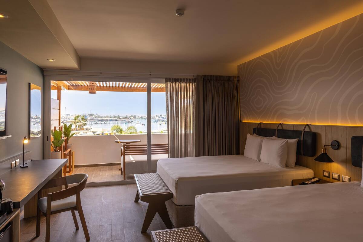 A hotel room with two beds, a desk, and a balcony with a view.