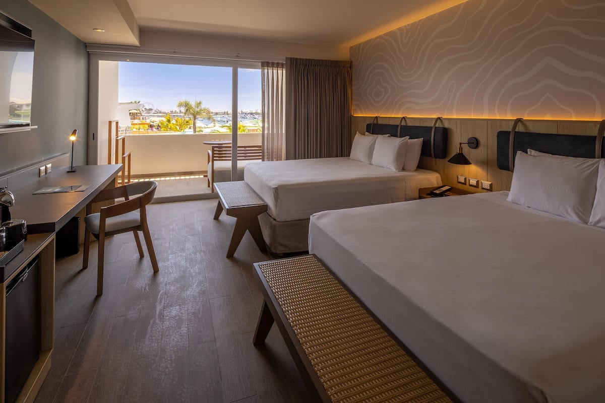 A hotel room with two beds, a desk, and a chair. There is a balcony with a view of the city.
