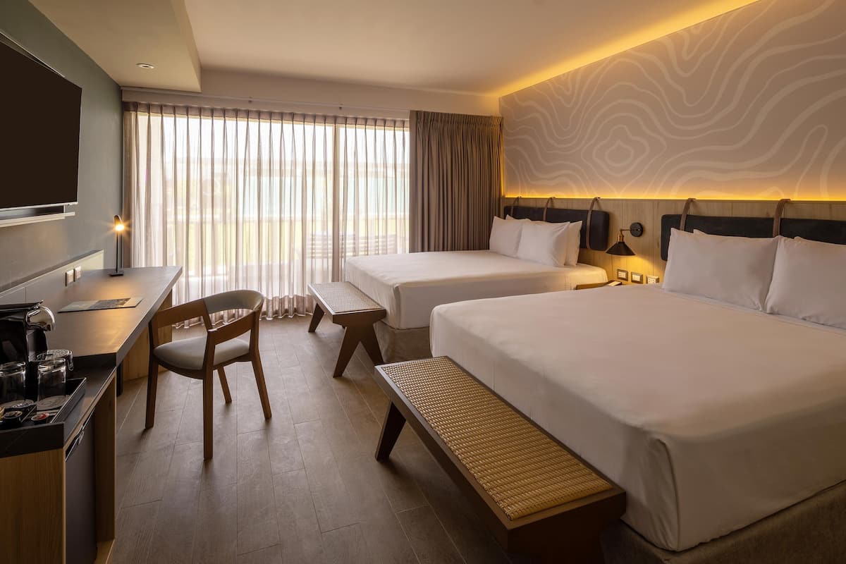A hotel room with two beds, a desk, a chair, a bench, a TV, and a window with curtains.