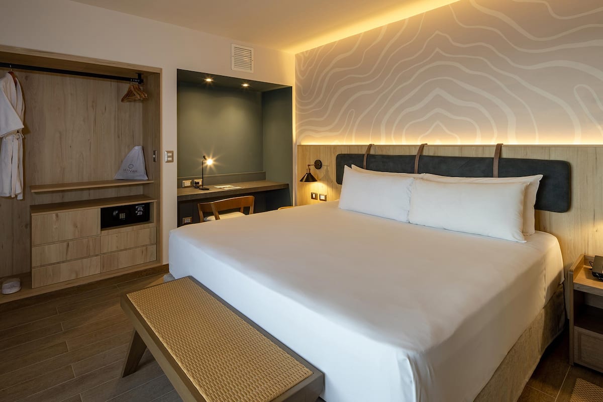 A hotel room with a large bed, a wooden wardrobe, a desk with a chair, and a bench.