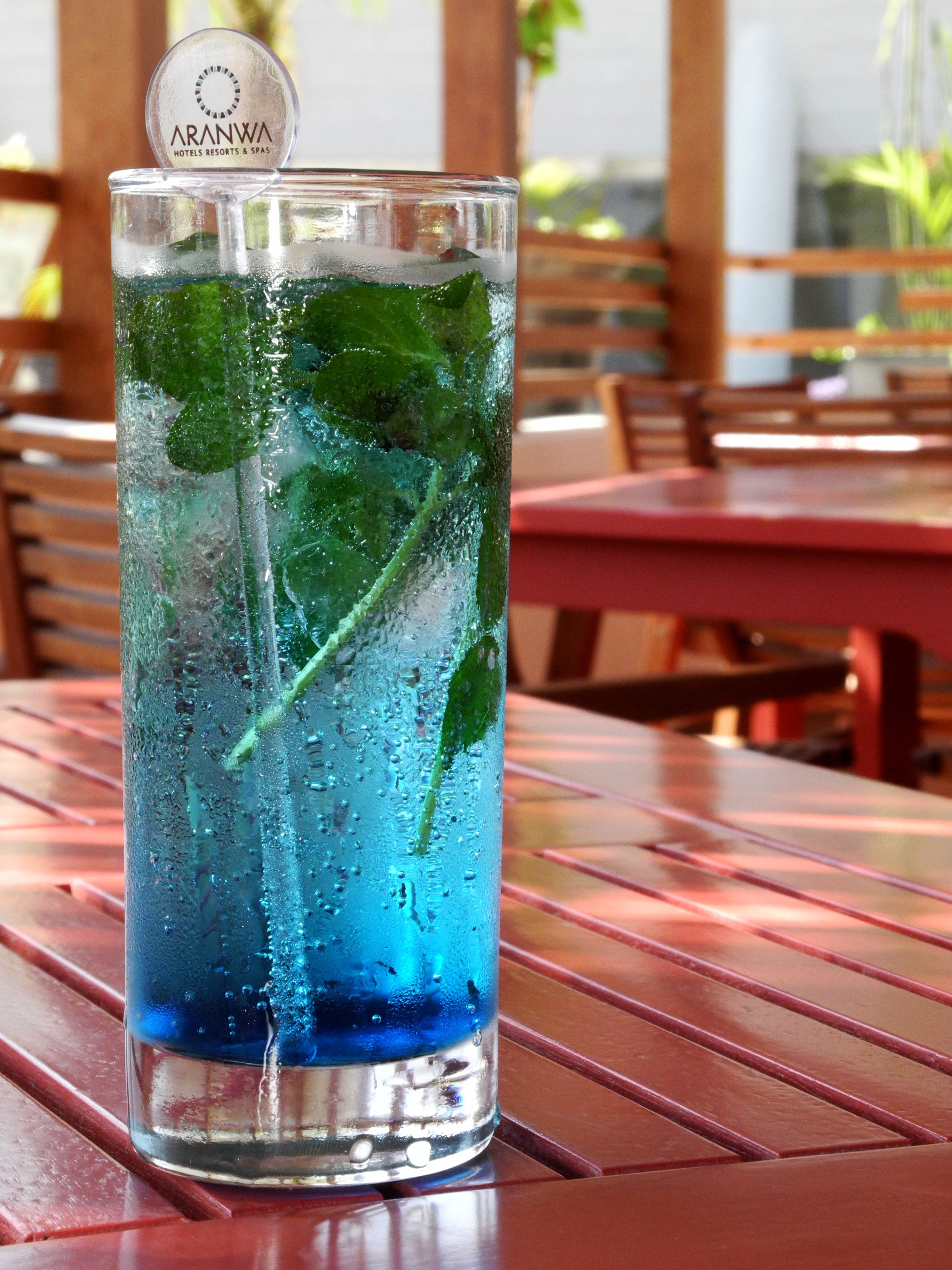A glass of blue beverage with mint leaves, labeled 'ARANWA HOTELS RESORTS & SPAS', on a wooden table.
