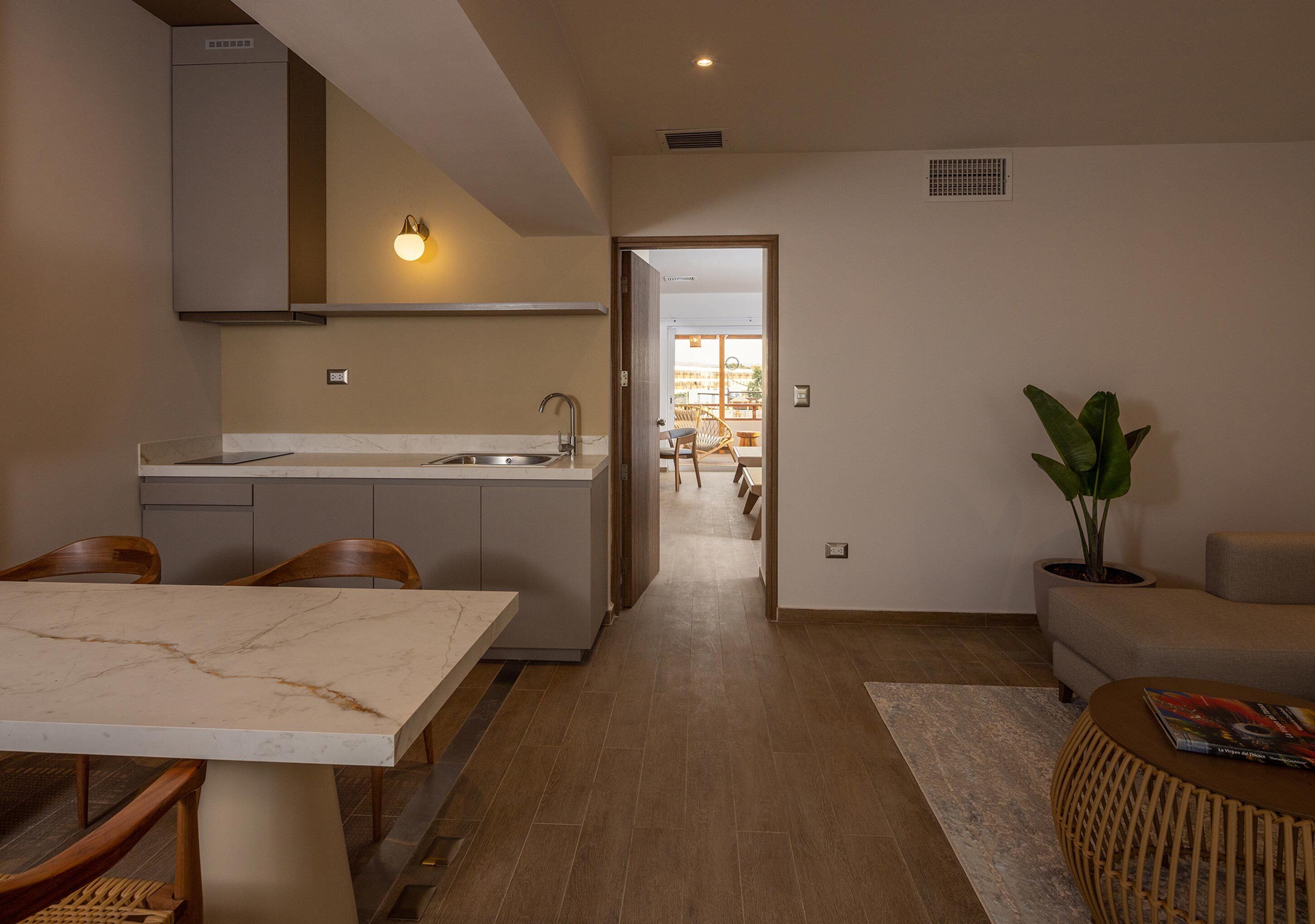 A modern kitchen with a marble countertop, wooden chairs, and a dining table. There is a sofa and a potted plant in the living area.
