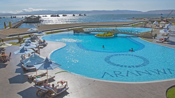 A large swimming pool with a bridge, surrounded by lounge chairs and umbrellas, with a view of the sea and mountains in the background. - Aranwa Paracas Resort & Spa (Paracas)