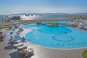 Una amplia piscina con un puente, rodeada de sillones de playa y sombrillas, con vista al mar y montañas al fondo.