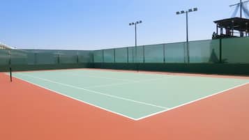 An indoor tennis court with a green playing surface, surrounded by a green fence, and equipped with overhead lighting.