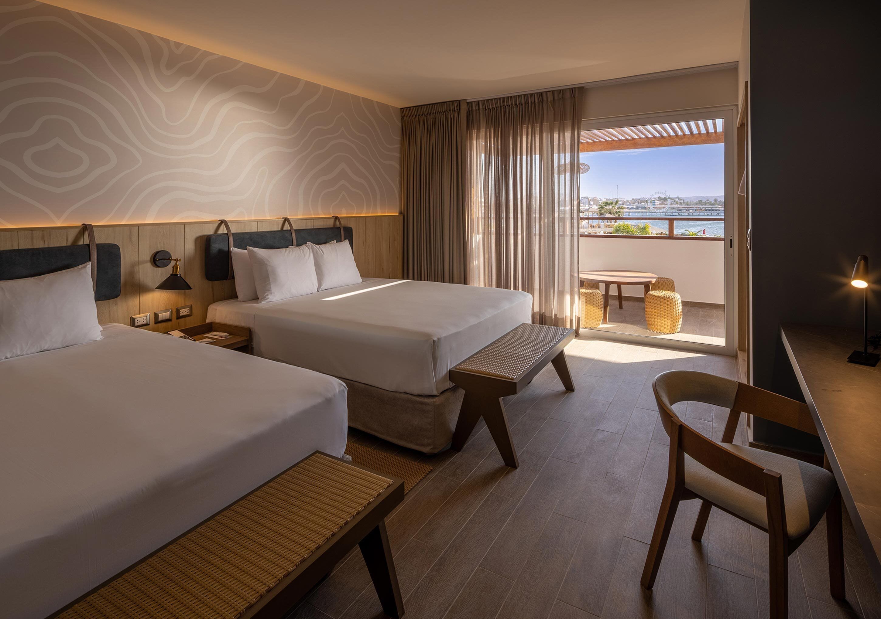 A hotel room with two beds, a small table, a chair, and a balcony with a view.