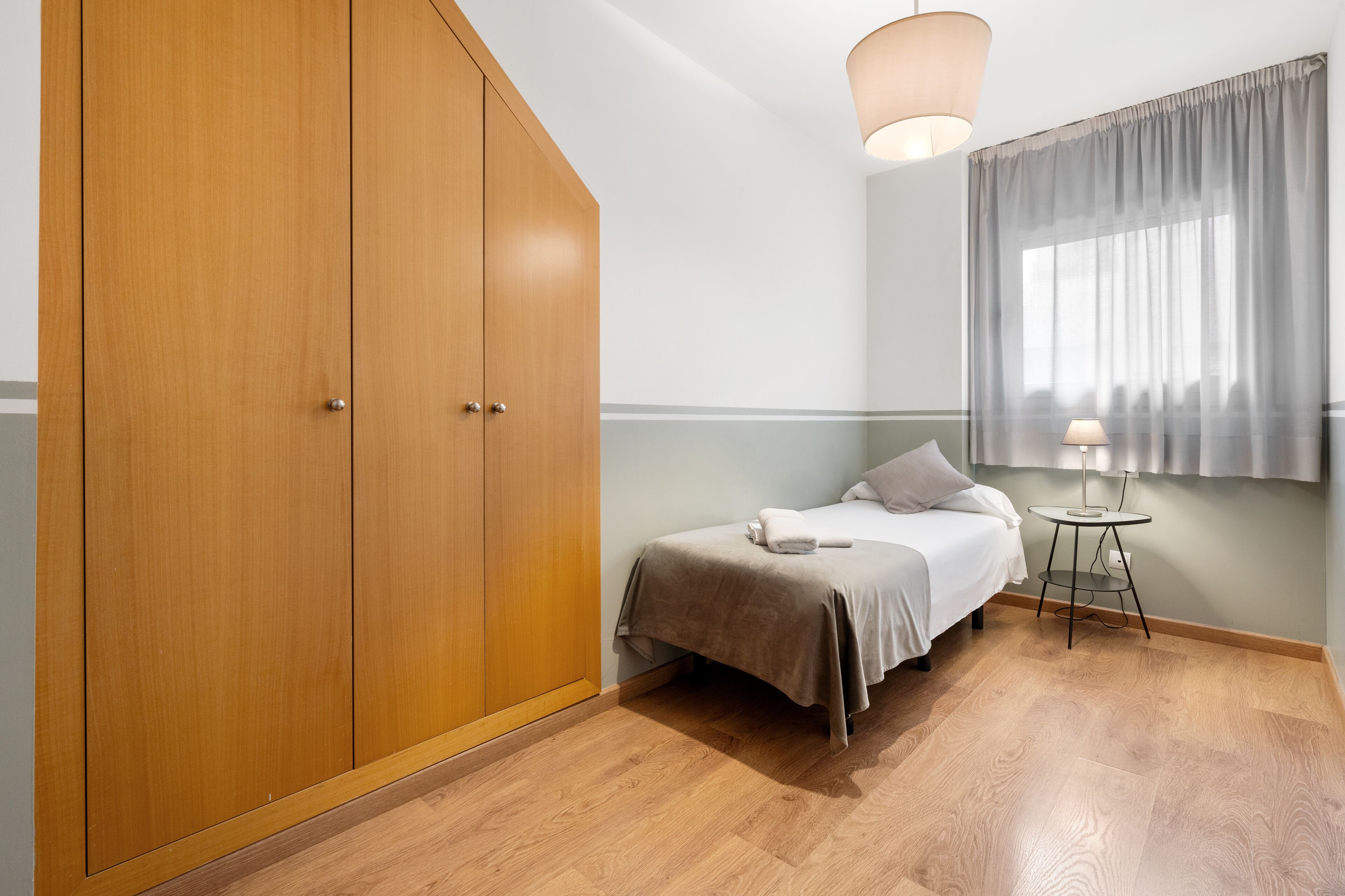 Une chambre à coucher avec un lit, une armoire, une petite table avec une lampe et une fenêtre avec des rideaux.