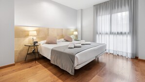 Een moderne hotelkamer met een groot bed, nachtlampjes, een tafeltje en stadszicht door dunne gordijnen.
