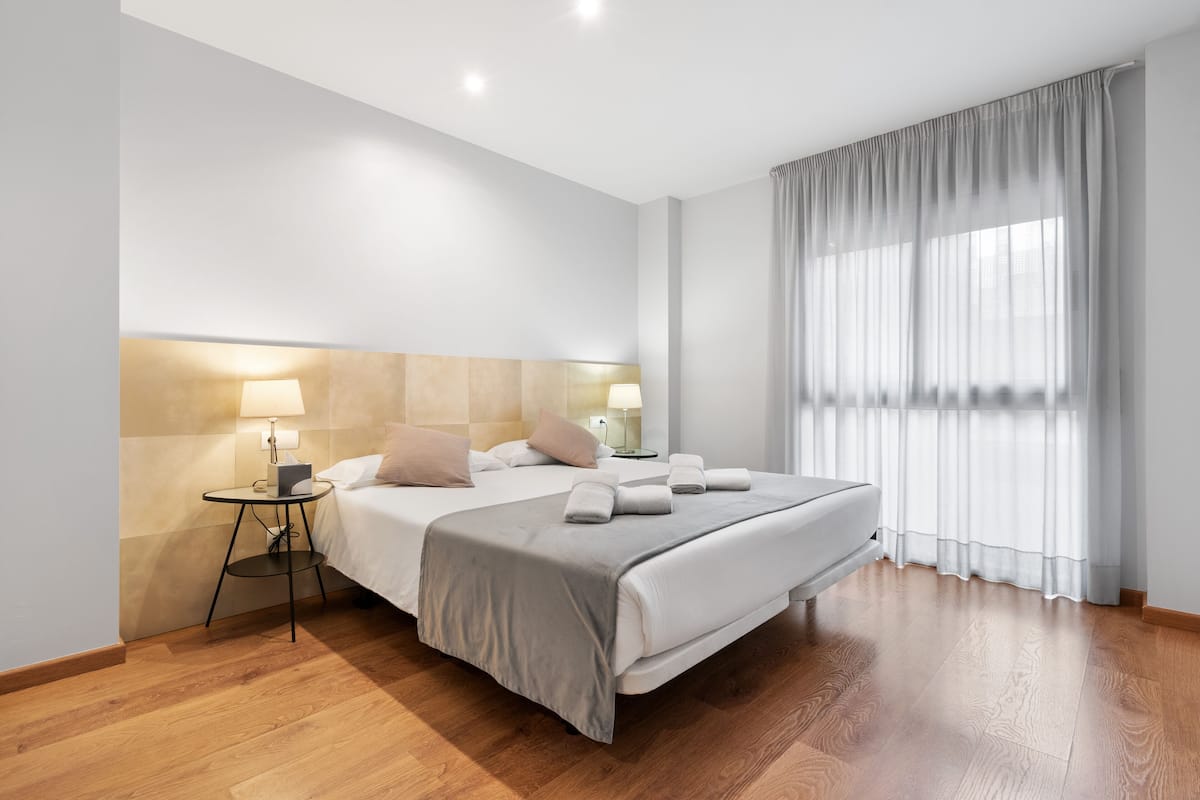 Habitación de hotel moderna con una cama grande, lámparas de noche, una mesita y vistas a la ciudad a través de cortinas translúcidas.