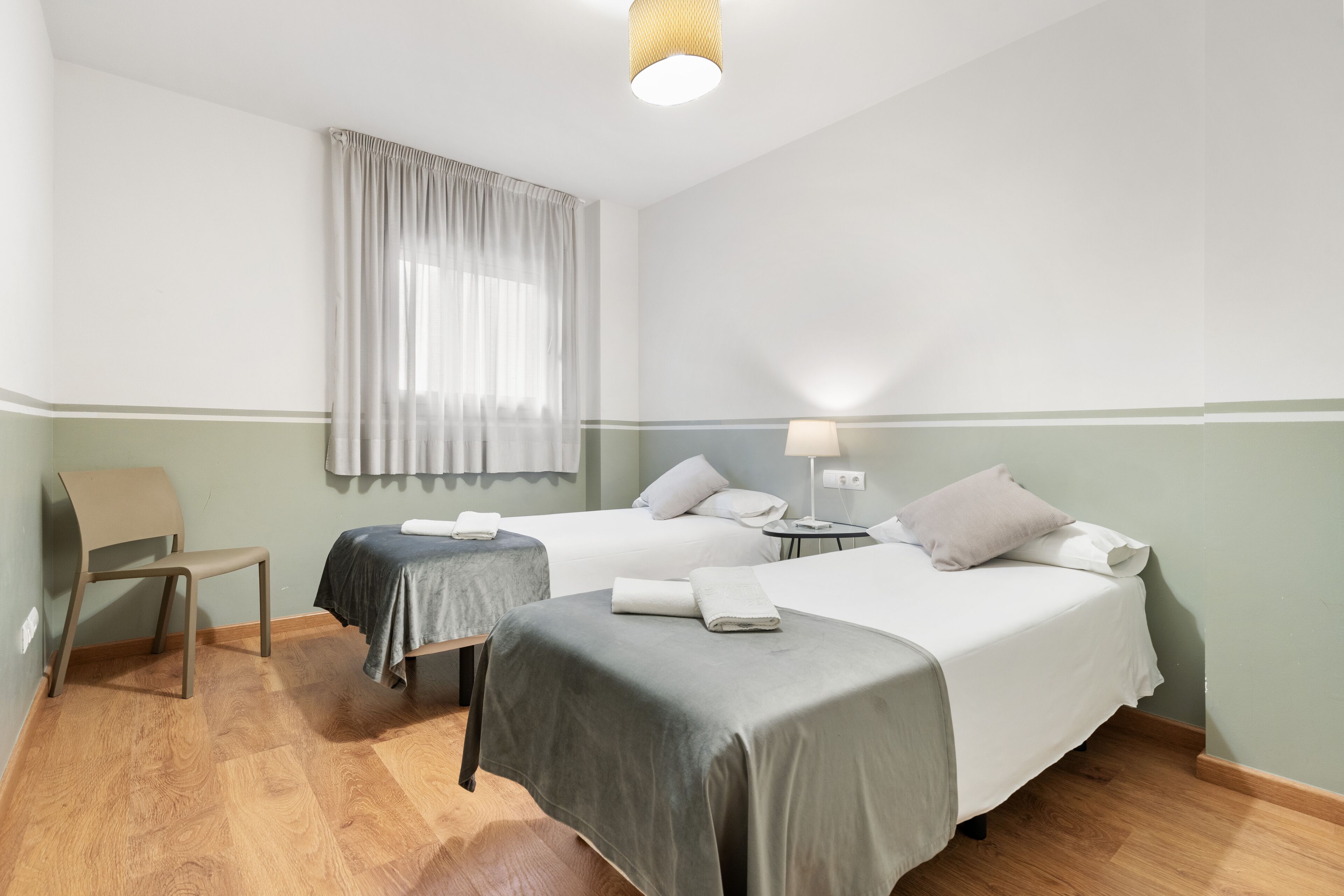 A hotel room with two beds, a chair, a small table, and a window with curtains.