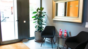 Een receptiegedeelte met een potplant, twee zwarte stoelen, een kleine tafel met brochures en een spiegel aan de muur.
