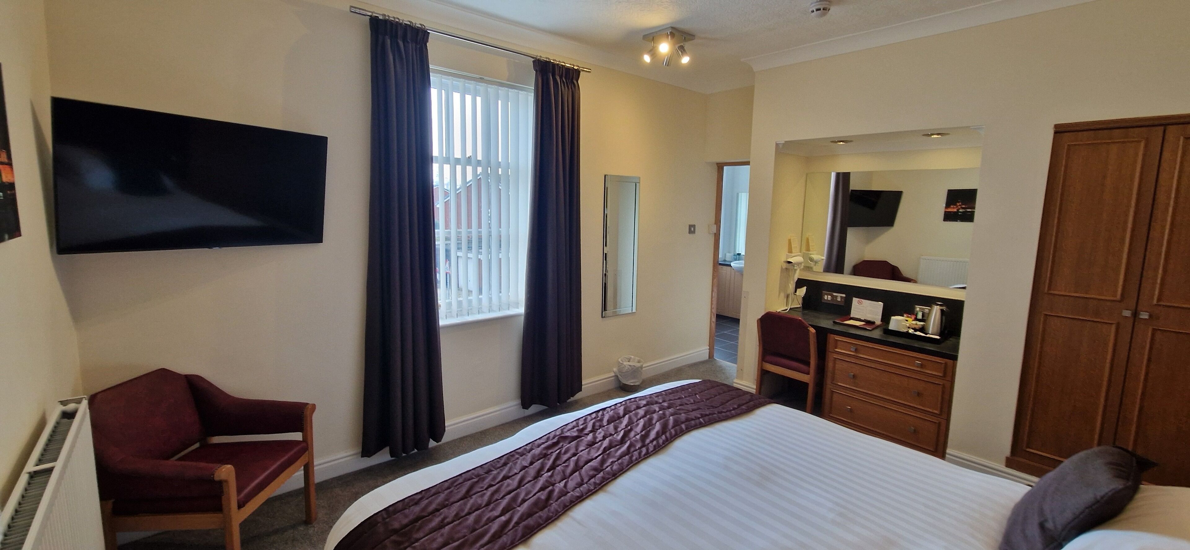 A hotel room with a bed, a desk with a mirror, a chair, a TV mounted on the wall, and a window with curtains.