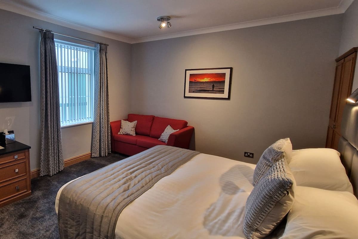 A hotel room with a bed, a red sofa, a TV, a window with curtains, and a framed picture on the wall.