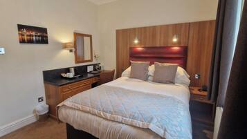 A hotel room with a large bed, a wooden headboard, a bedside table, a nightstand, a desk with a chair, a mirror, and a wall-mounted lamp.