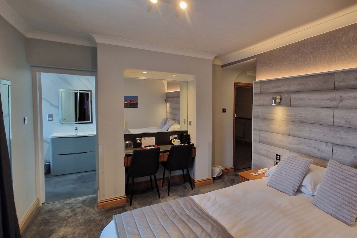 A hotel room with a bed, a desk with a chair, a mirror, and a wooden headboard.