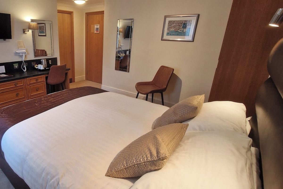A hotel room with a large bed, a desk with a chair, a mirror, a television, and a bathroom visible through an open door.
