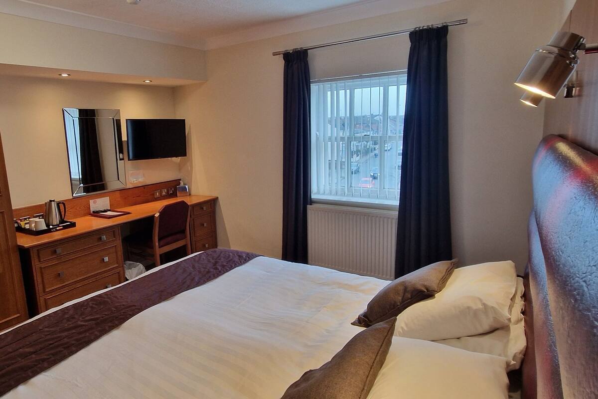 A hotel room with a large bed, a wooden wardrobe, a desk with a chair, a flat-screen TV, and a window with curtains.