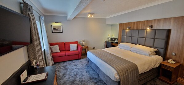 A hotel room with a bed, a red sofa, a TV, a bedside table with a lamp, and a small table with a book and remote control.