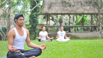 A group of people meditating on a grassy area with a thatched-roof structure in the background.