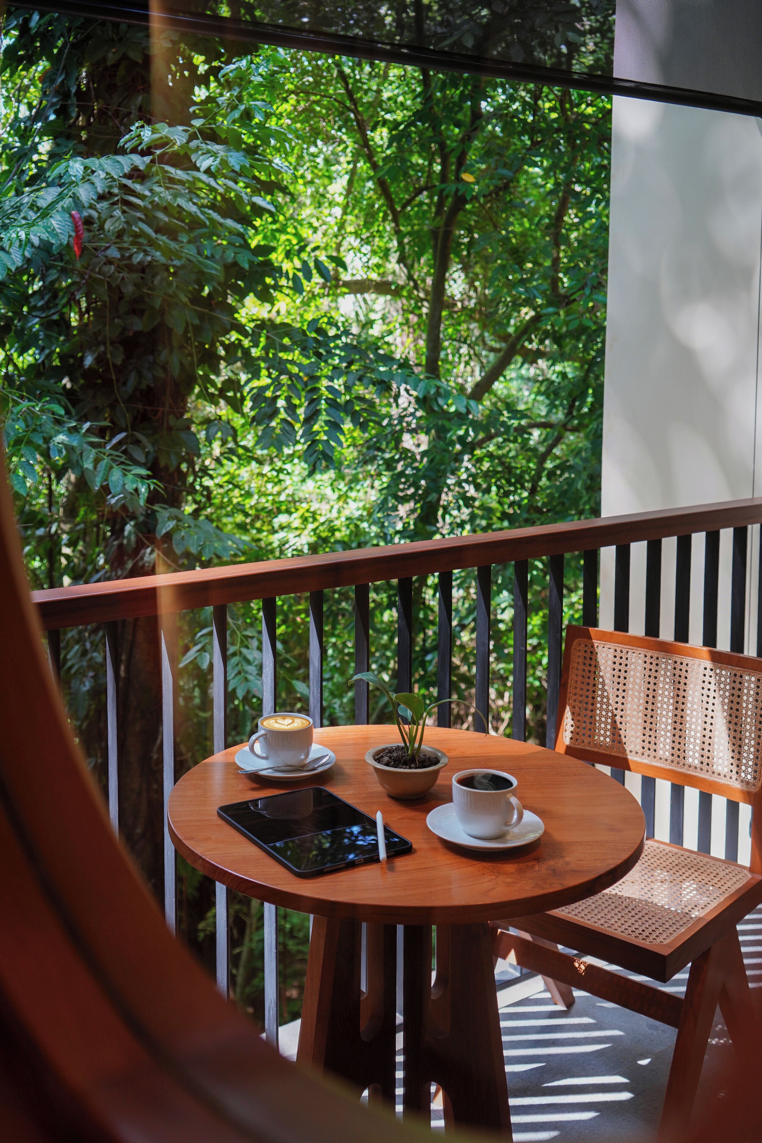 A wooden table with a cup of coffee, a smartphone, and a small potted plant on a balcony with a view of dense greenery.