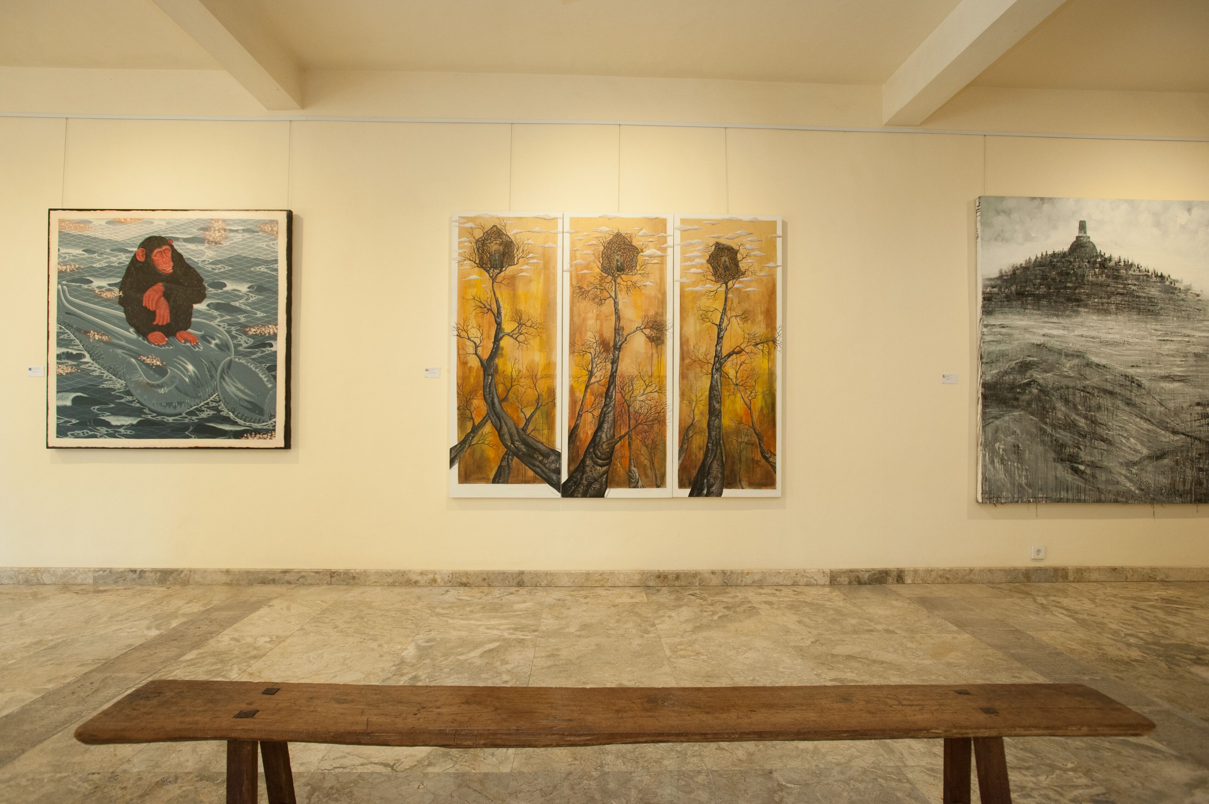 A gallery with three paintings on the wall and a wooden bench in the foreground.