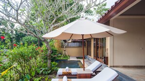 Zona junto a la piscina con sillones de descanso y una sombrilla, rodeada de abundante vegetación y una piscina pequeña.