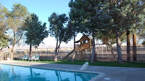 Un espace piscine avec une terrasse en bois, une niche pour chien et une cour clôturée avec des arbres.