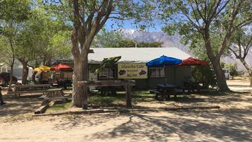 Un café al aire libre con mesas tipo picnic y sombrillas, rodeado de árboles y con una montaña al fondo.