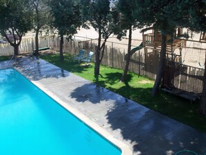 A swimming pool with a wooden deck and a fenced area with trees and a swing set.