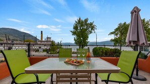 An outdoor dining area with a table and chairs, a bottle of wine, and a view of a body of water and mountains. - Casa Grande Inn (Penticton)